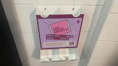 A plastic dispenser of free period products. The dispenser has a large sticker on the front that says 'free period products take what you need, when you need it'