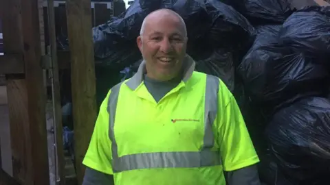 Dave Callaghan A man wearing a hi-vis jacket standing on front of a pile of black bin bags filled with rubbish.