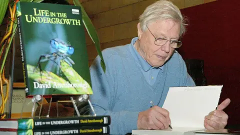 Getty Images A man white white hair and glasses signs a book with a pen, with the book on display to the right of him showing an insect on the front page and "Life In The Undergrowth" as the title.