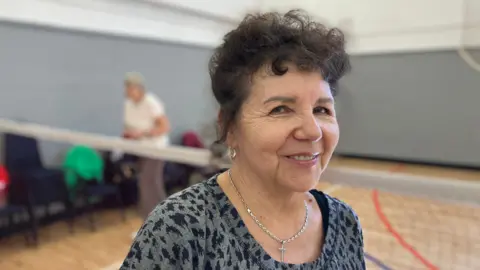 Shariqua Ahmed/BBC Luda, a woman, with curly short brown hair. She is wearing a grey printed top and is smiling for the camera.