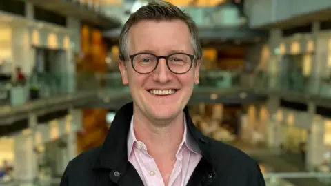 BBC News Alex Green smiling with offices down  him