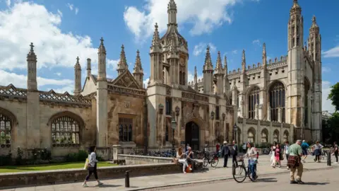 King's College in Cambridge on a sunny day. There are people walking on the pavement and public road in front of the gate and its chapel. 