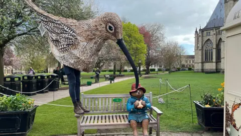 BBC/Richard Fox A man is in York's Deans Park, dressed in a large speckled brown curlew costume featuring a long beak, with his legs visible underneath. He's wearing navy trousers and brown leather boots. He's standing on the arm of a bench, leaning toward a statue of Paddington Bear eating a sandwich, as if trying to take it.