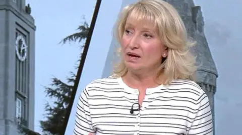 Sian Astley, wearing a white top with thin black stripes, stands in a TV studio