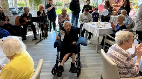 BBC/TomWalker Marjorie sits in a wheelchair in the centre of a community room at the care home in which she lives. People can be seen clapping their hands on tables around her. Birthday decorations including balloons can be seen across the room. 