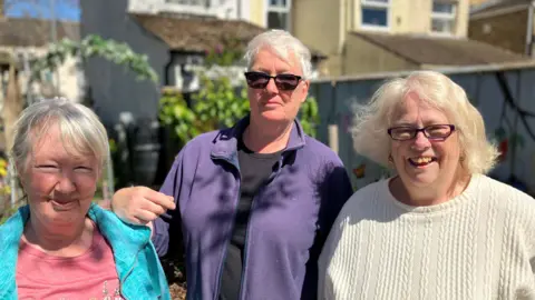 Shariqua Ahmed/BBC Pippa, a woman with short white hair, wearing a blue fleece and a pink top, Mo with short white hair - wearing a purple fleece and black glasses and Erica with short white hair -wearing a white jumper- all smiling for the camera.