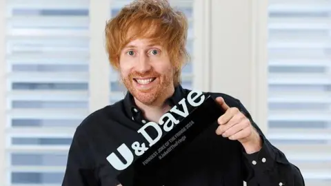 PINPEP/Robert Perry Comedian Mark Simmons, with messy ginger hair wearing black shirt holding Joke of the Fringe trophy at an angle. 