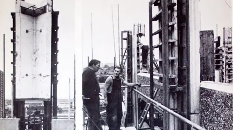 Constantine Gras Black and white photo showing one of the walls of the tower with two men by the side of the scaffolding talking to each other.
