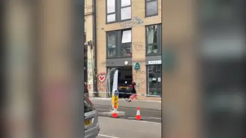 A black BMW that has crashed into a pizza restaurant. There is police tape and traffic cones cordoning off the scene.