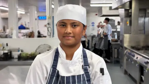 Marize Sabapathy in chef's whites and hat and a blue and white apron in a kitchen