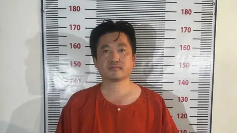 In an orange prison-style uniform, Cambodian businessman Kuong Li stands facing the camera in front of a height-measurement chart