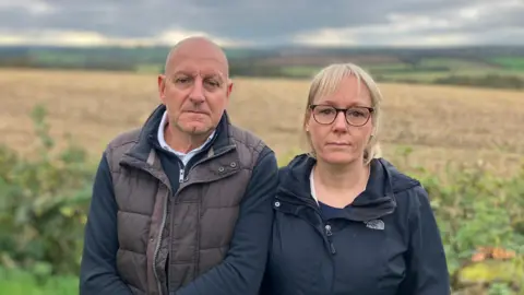 BBC Stephen and Sharon Boulton stand in front of fields in the open countryside.
Stephen Boulton is bald and is wearing a dark body warmer over a long-sleeve shirt.
Sharon Boulton is wearing a dark jacket and has blonde hair and spectacles.
The sky is cloudy and grey.