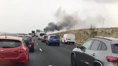 A queue of stationary traffic. Black smoke can be seen rising in the distance