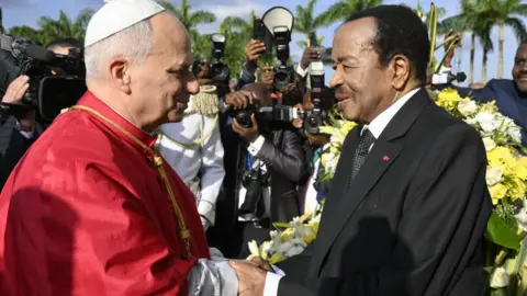 Pope Leo in a red cape shakes the hand of President Paul Biya in a black suit with a white dotted tie. Behind them are banks of photographers and a floral arrangement.