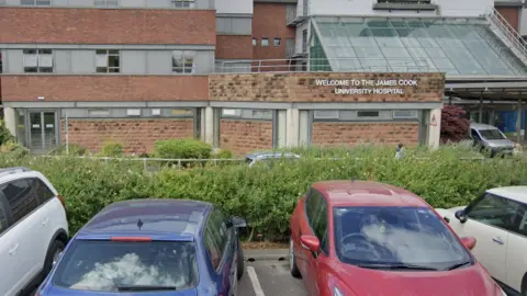 A row of cars parked in front of a hedge outside the entrance to the James Cook University Hospital.