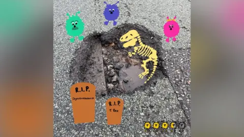 A photo of a pothole in a road that has drawings on top of it. The drawings depict a dinosaur skeleton in the pothole, with two grave stones below it. Above the pothole are three fuzzy creatures - one pink, one blue and one green