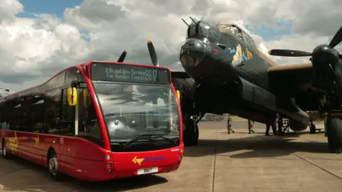 A red bus parked next to Just Jane - a World War Two Avro Lancaster bomber. The sky is cloudy.