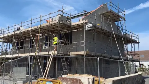 BBC A man in a luminous vest standing on scaffolding among a brickwork house frame, surrounded by a fence. 