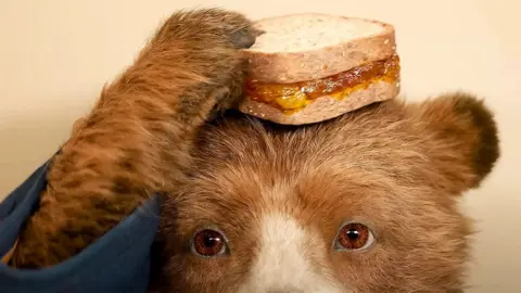 Still from the film Paddington 3 showing Paddington reaching for a marmalade sandwich that is balanced on his head