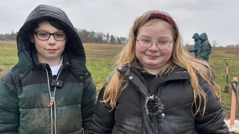 Henry and Rebecca smile at the camera. Henry has a green and black winter coat on and his hood on, and black square-framed glasses. Rebecca has long blonde hair, round glasses and a red headband. She wears a black coat. Both of them are muddy, and the field behind them is clearly wet. 