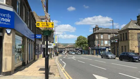 Aisha Iqbal/BBC A town centre high street. A yellow sign points to the Five Rise Quarter, identifying the town as Bingley. Shops line the street and there's light to busy traffic and pedestrian activity.
