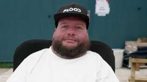 Karl Flood looking directly to the camera. He is sat in a wheelchair and has a white T-shirt on and black hat.