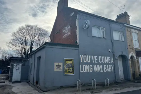 An end terrace house painted grey, with signage reading "The New Adelphi Club" and large white lettering on the wall saying "You’ve come a long way, baby". A bicycle stand and an arched doorway are situated on the front of the building. Trees and a cloudy sky are visible in the background.