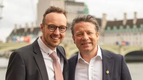 Adam Dance and Jamie Oliver stand side by side, smiling at the camera. They are both wearing white shirts and suit jackets. Dance is wearing a light pink, patterned tie. The background is blurred but they appear to be standing in front of government buildings.