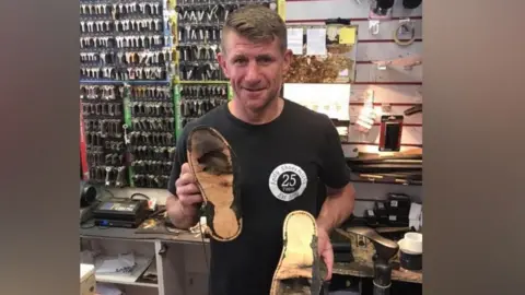 Andy Smith A man wearing a black T-shirt and holding a pair of damaged shoes in a workshop with racks of keys in the background.