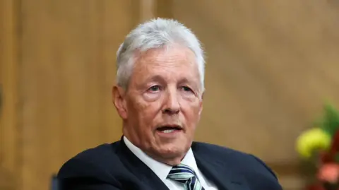 Pater Robinson. He has short white hair, wearing a navy blazer, white shirt and green and navy tie.