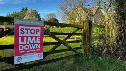 A Stop Lime Down sign strapped to a traditional wooden five-bar gate outside a green paddock with houses around.