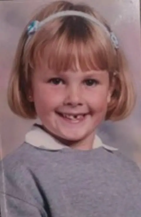 Leonie Hughes A picture of a roughly five-year-old blonde-haired girl smiling, with a gap between her front teeth. She wears a grey jumper a white shirt.
