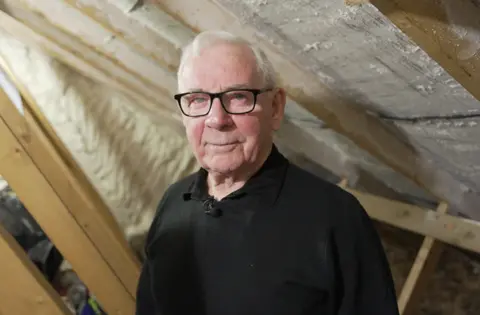An older man with grey hair and wearing glasses and a black jumper is standing under insulation in his home's roof space