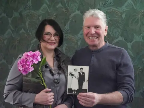 BBC Iryna and Serhiy photographed in 2026, looking at the camera and smiling. Iryna has chin-length dark hair and glasses and is holding a pink flower. Serhiy has grey hair and is holding up a photo of them side-by-side at their wedding. 