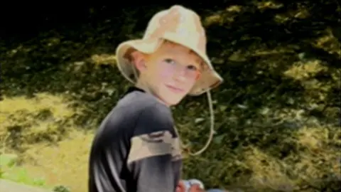 Zac Roe looks over his shoulder at the camera. He is wearing a dark top and a floppy summer hat. The background shows tree shadows on grass.