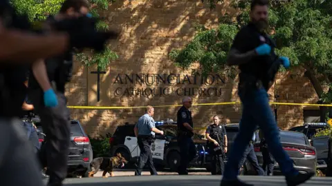 Getty Images Police officers, one leading a police dog, and parked police cars are seen near a sign reading Annunciation Catholic Church on a stone wall of the church. There is a line of yellow police tape along the building. 