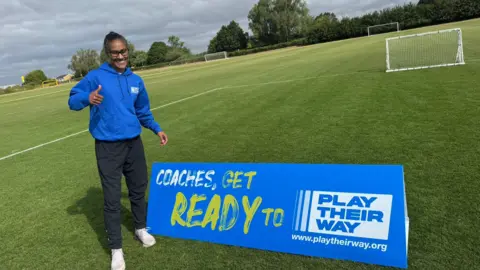 A smiling Rachel Yankey next to a blue sing on the pitch reading "Coaches, get ready to play their way", with the campaign's website listed below. She is giving the thumbs up. It's cloudy.