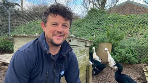 Dave Rich, wearing a blue zip up jumper, with black hair and beard. He is sat to the left of the image. He is looking directly at the camera. Behind him is a small wooden hut. There are five ducks walking. Behind them is a metal fence and green bushes.