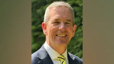 East of England Ambulance Service Trust A headshot of a man with short grey hair who is wearing a navy suit and a white shirt with a yellow and navy striped tie.