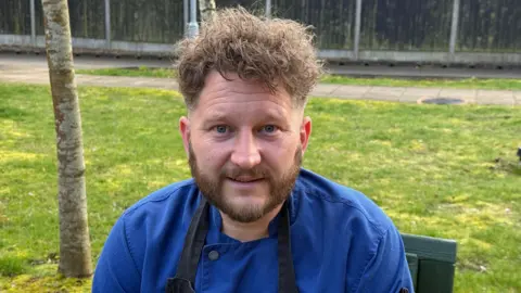 A man wearing a blue top and a black apron is sitting in green wooden chair outdoors. He is looking directly at the camera. There is a lawn, a pathway and some fence panels behind him.