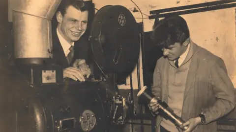 Ricky Ford Two men next to an old-fashioned film reel projector. Ricky,a young man, is on the right, wearing overalls and a shirt and tie. An older man is smiling left, and leaning over the projector