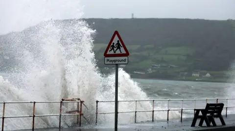 Image of waves crashing against a coastal path. The backdrop behind the waves is a green hill with trees and houses.