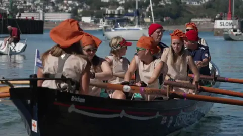 Women wearing cream coloured aprons and orange mob caps are rowing the gig with several men from the lifeboat in the crew too. They are in a harbour.
