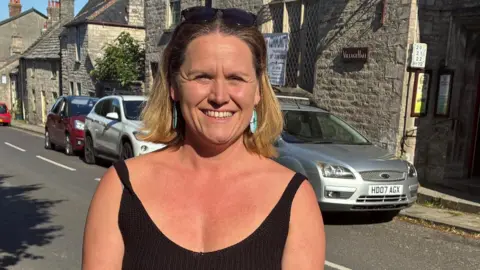 A smiling Lauren Malone stands on the pavement opposite the school. She is wearing a black vest top and has shoulder length blonde hair pushed back by sunglasses resting on top of her head.