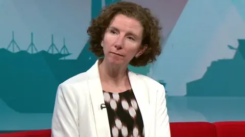 Dame Anneliese Dodds has medium length brown hair, and is wearing a white blazer over a black and white top. She is sitting on a red sofa with a red and blue wall of graphics behind him.