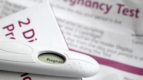 A positive pregnancy test lies on top of the test box which is white with magenta writing, the instructions for the kit are blurred out in the background.