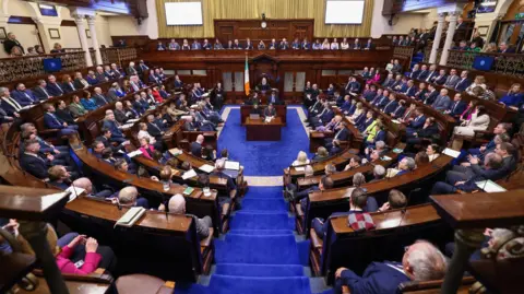 PA Media A full sitting of the Dail, the Irish Parliament from January 2025. The chamber has curved and straight wooden benches, a blue carpet and a podium in the back middle of the picture.