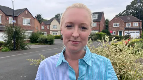 Natalie Bell/BBC A woman with blonde hair, pulled back into a ponytail, wears a blue striped shirt and pearl earrings. She is stood outside houses on a quiet residential street.