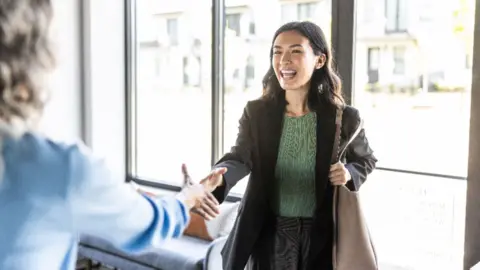 g woman with long dark hair wearing a blazer over a green top, shaking someone's hand.
