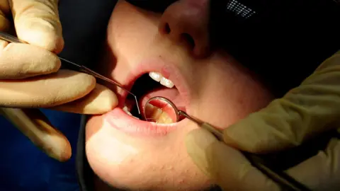 A close-up image of a dentist using a scaling tool and mirror to look at a child's teeth. The dentist is wearing gloves.